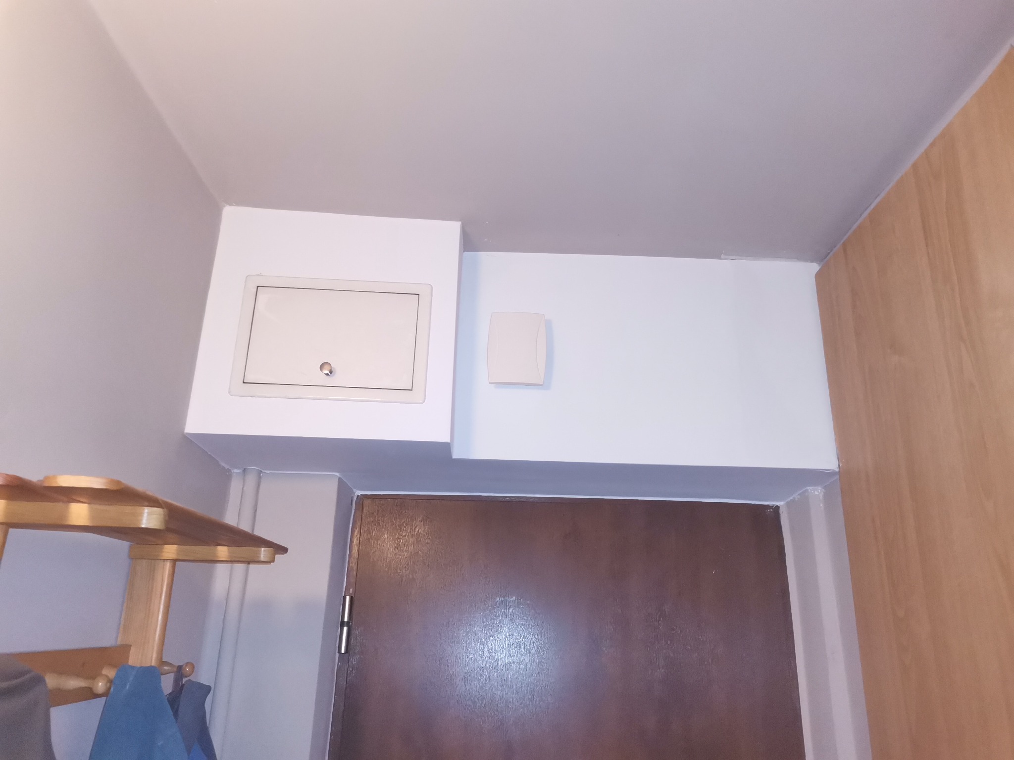 Biała klapa rewizyjna na suficie, obok czujnik, widoczna brązowa futryna drzwi i drewniany wieszak na ubrania.