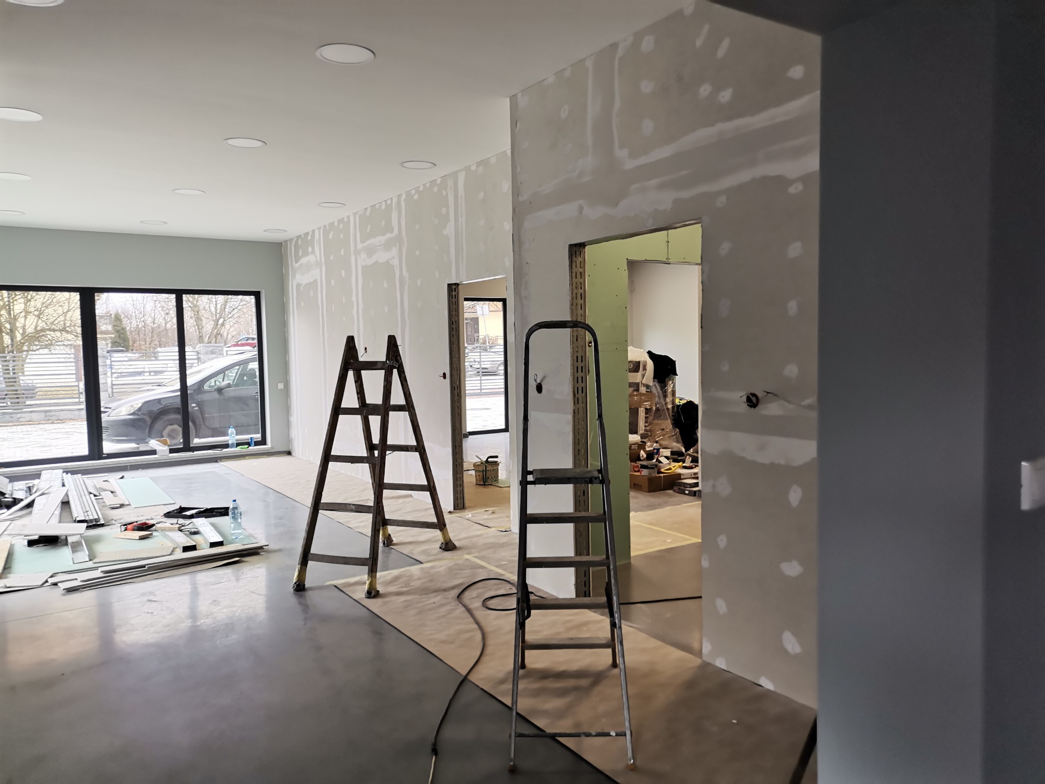 Wnętrze w trakcie wykańczania: ściany pokryte płytami gipsowo-kartonowymi, widoczne łączenia szpachlowane, dwie drabiny, narzędzia i materiały budowlane na podłodze, duże okno z widokiem na zewnątrz.
