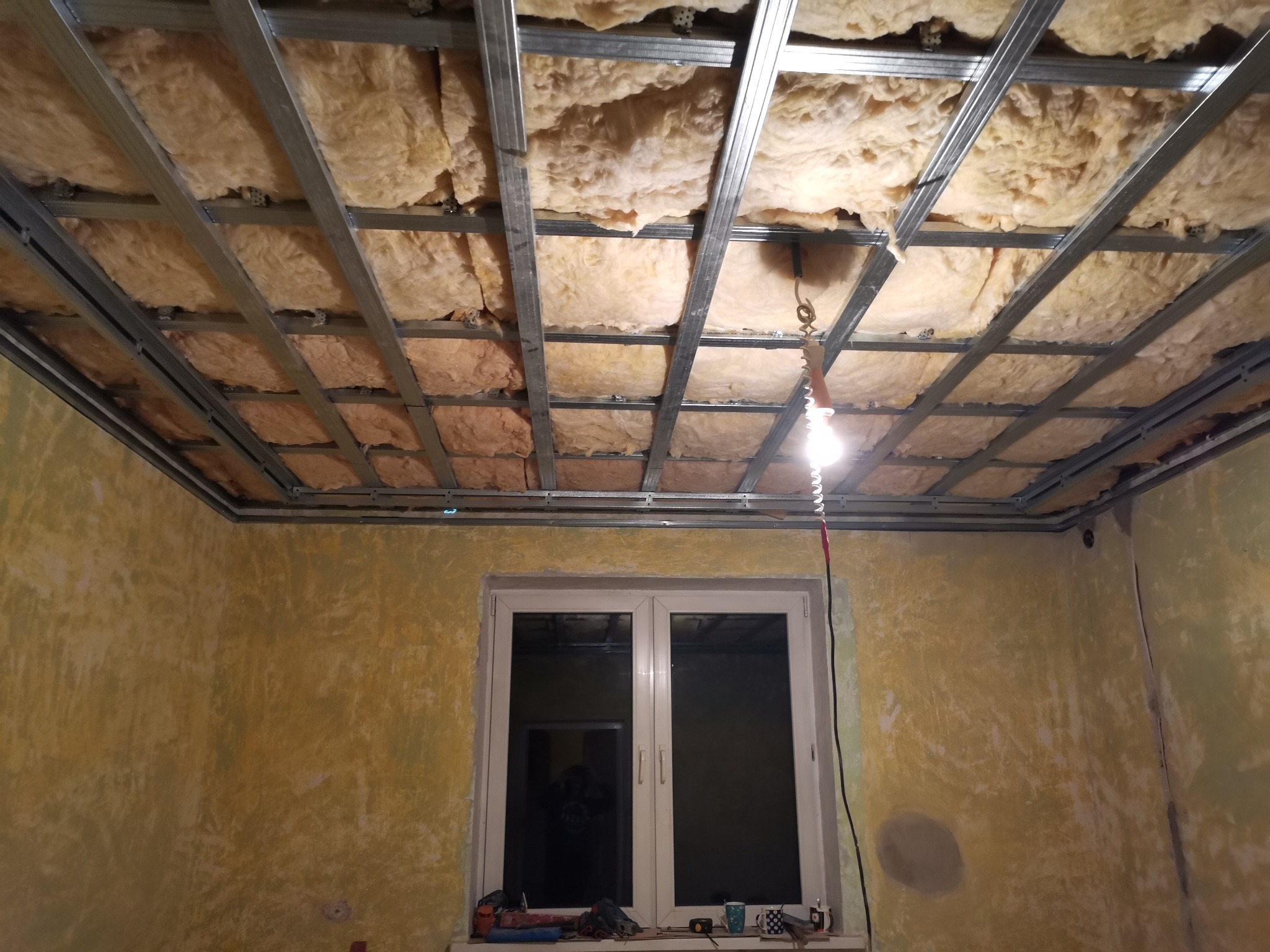 Montaż metalowego stelażu sufitowego z wypełnieniem z wełny mineralnej, widok na okno i fragment ściany z nierówną fakturą, oświetlone wiszącą żarówką.