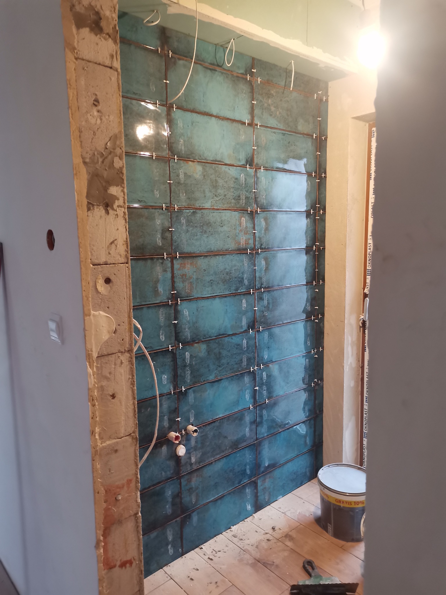 Ściana w trakcie układania turkusowych płytek, widoczne krzyżyki dystansowe, wystające rury instalacji hydraulicznej, surowe wykończenie ościeżnicy i wiadro z zaprawą.