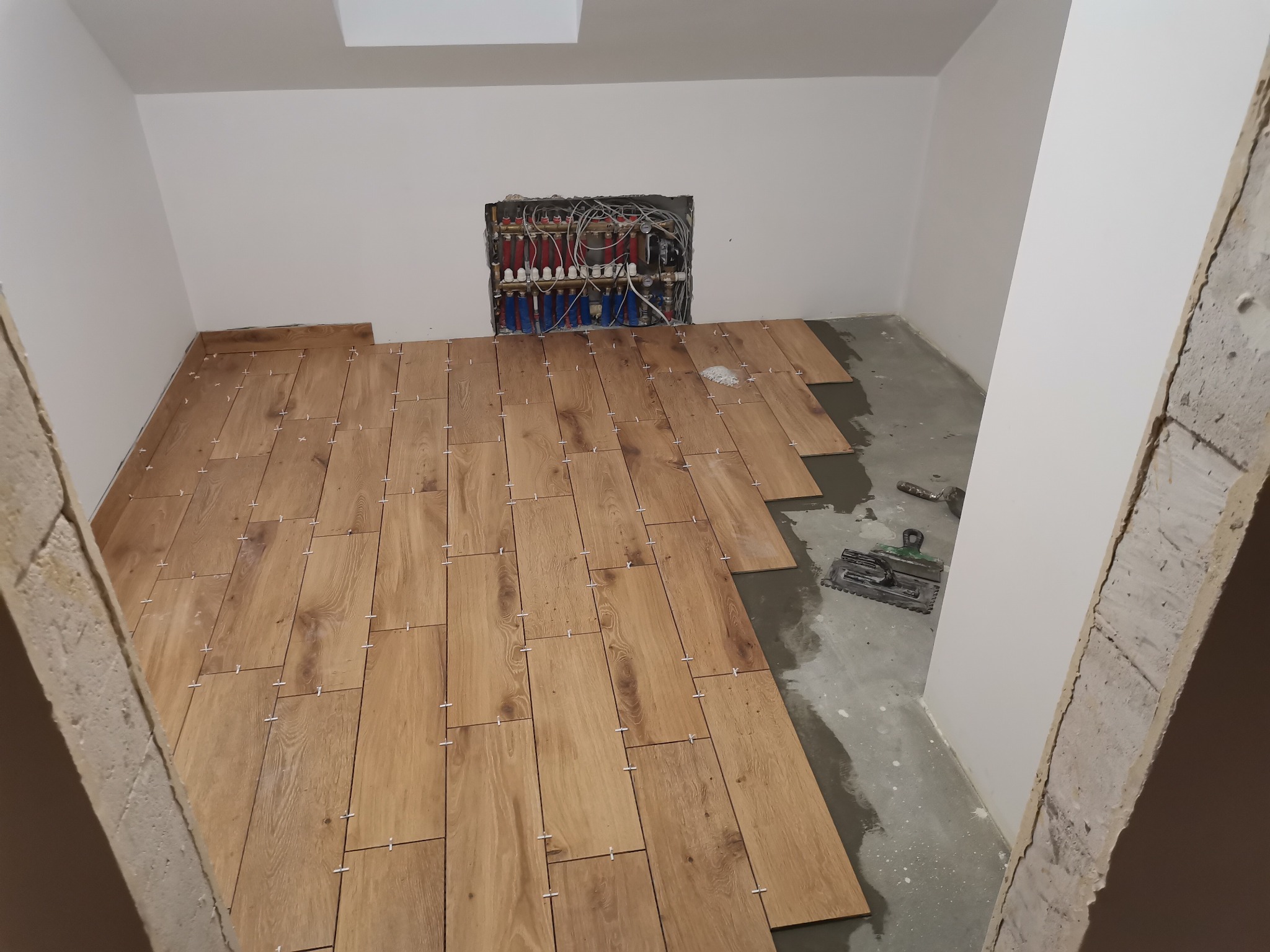 Układanie drewnopodobnych płytek podłogowych z użyciem krzyżyków dystansowych, widoczna instalacja grzewcza w ścianie i narzędzia glazurnicze.