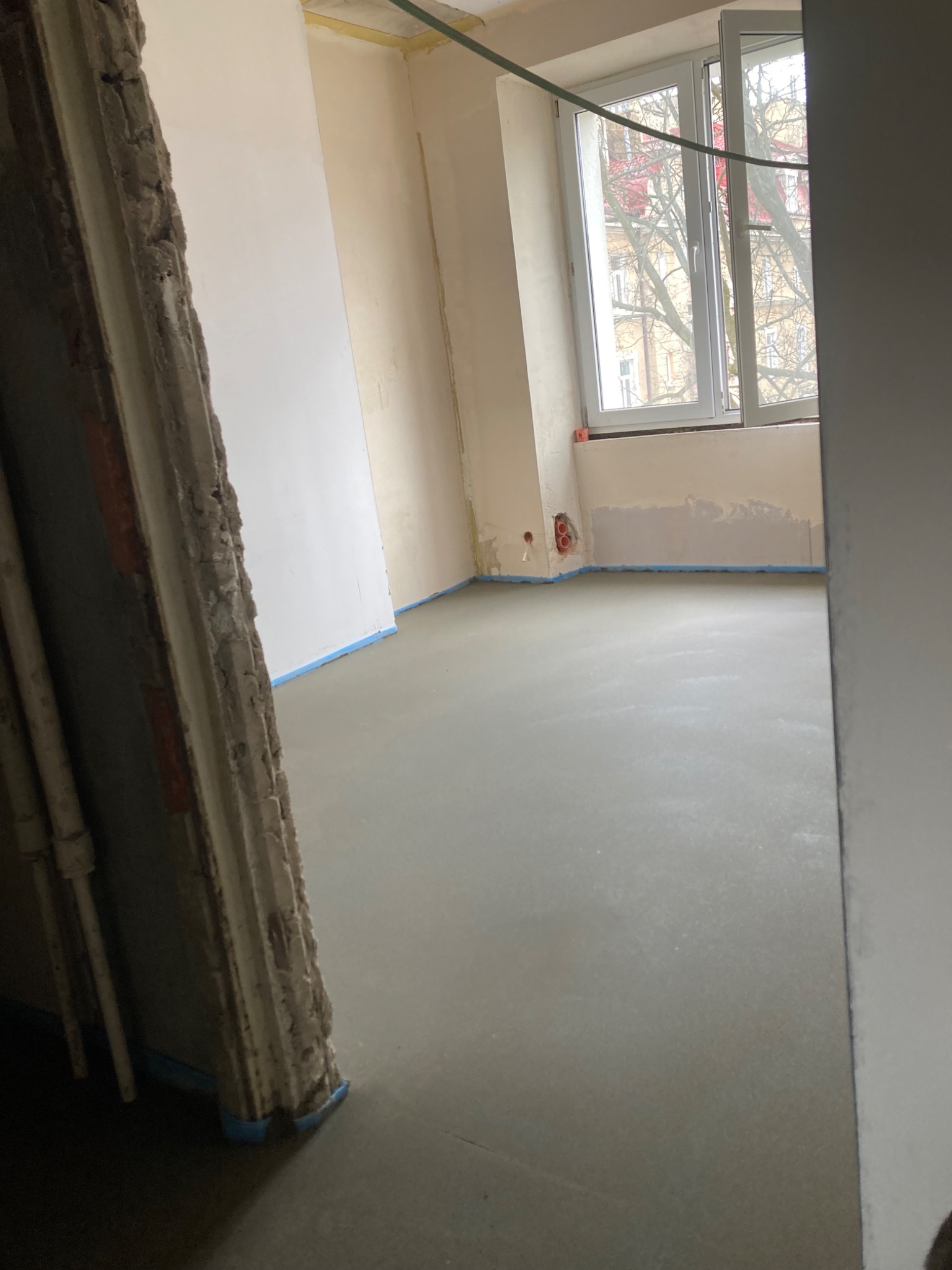 Świeżo wylana, gładka, szara posadzka w pomieszczeniu w trakcie remontu, zabezpieczone ściany taśmą malarską, widoczne okno z widokiem na drzewa.