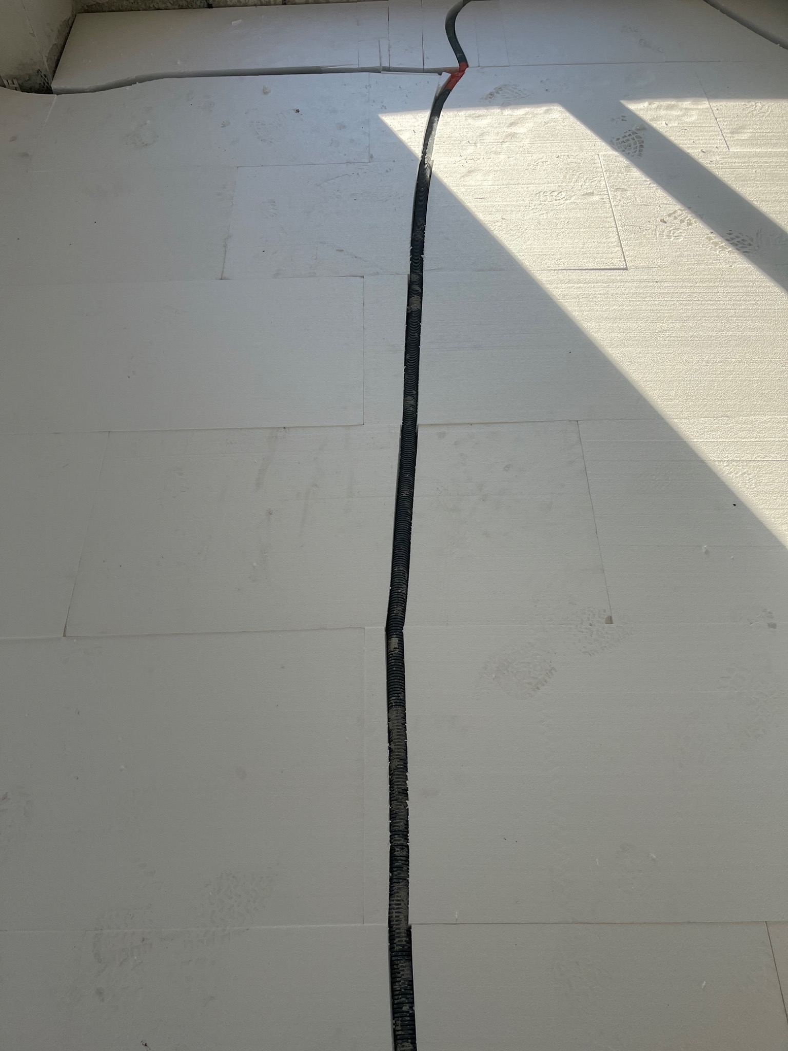 Białe płyty izolacyjne pod wylewkę podłogową z czarnym przewodem w peszlu ułożonym w szczelinie, widoczne ślady butów i cień.