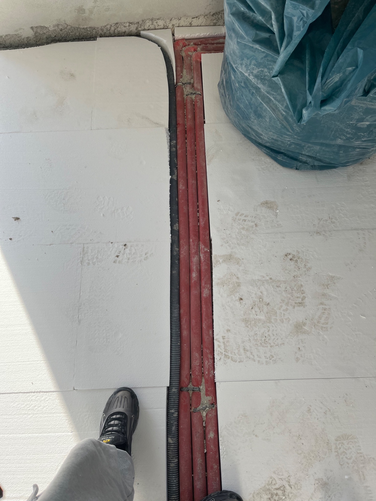 Białe płyty izolacyjne pod podłogę, czerwone rury grzewcze i czarna rura karbowana wzdłuż ściany, widoczny fragment buta i worek na śmieci w rogu pomieszczenia.