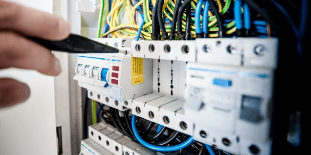 Instalacja elektryczna: Ręka montera przykręca śrubokrętem bezpiecznik w rozdzielnicy, widoczne kable w peszlach.