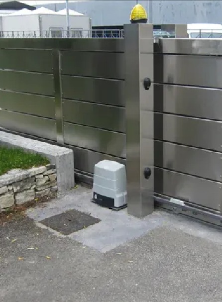 Automatyczna brama przesuwna ze stali nierdzewnej z żółtą lampką ostrzegawczą na słupku i napędem elektrycznym, widoczne elementy mechaniczne i betonowe obrzeże.