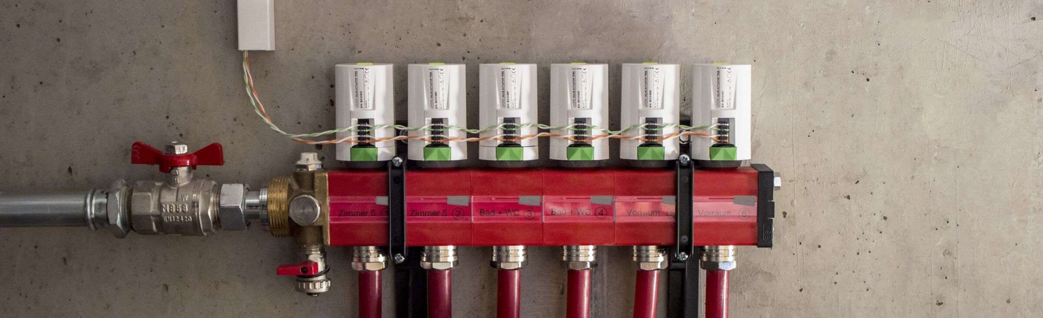 Rozdzielacz ogrzewania podłogowego z zaworami termostatycznymi i przewodami zainstalowany na surowej betonowej ścianie. Widoczne rury doprowadzające wodę, etykiety z nazwami pomieszczeń (Zimmer...