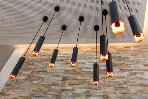 Nowoczesne oświetlenie sufitowe z cylindrycznymi lampami wiszącymi na cienkich przewodach, emitujące ciepłe światło, na tle ściany z kamienną okładziną.