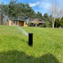 Automatyczny zraszacz trawnikowy w akcji, rozpylający wodę na soczystą, zieloną trawę, z altaną krytą strzechą i małym domkiem w tle, w słoneczny dzień.