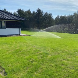 Systemy Nawadniania Kamil Kotlicki - Automatyczne nawadnianie trawnika w ogrodzie, zraszacz w akcji. W tle dom z ciemnym dachem i zielone drzewa. Błękitne niebo z delikatnymi chmurami.