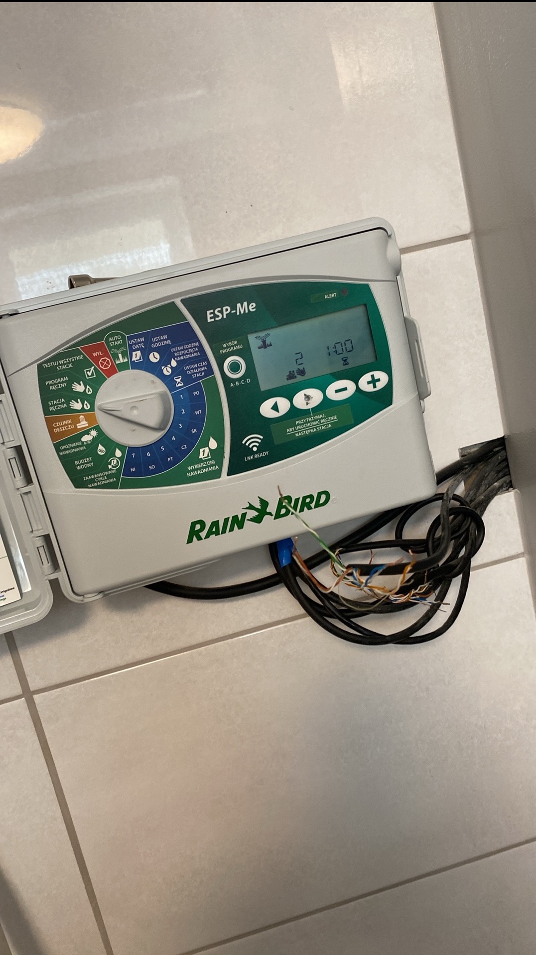 Sterownik nawadniania Rain Bird ESP-Me z otwartą obudową, widoczne kable i ustawienia. Detal montażu systemu nawadniania na tle płytek podłogowych.