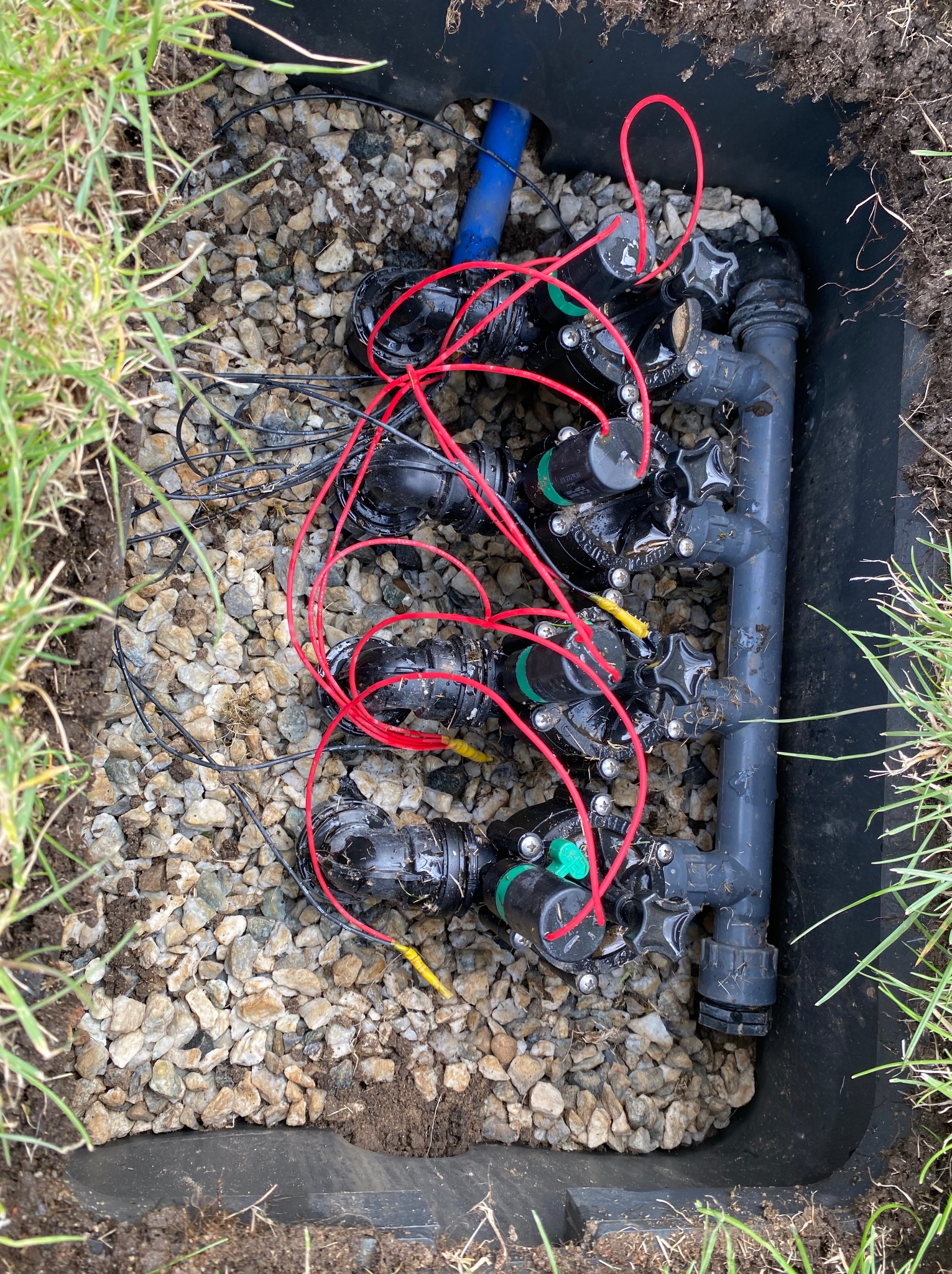 Skrzynka zaworowa z elektrozaworami i przewodami, wypełniona kamieniami, zainstalowana w ziemi, widok z góry. Element systemu nawadniania ogrodu.