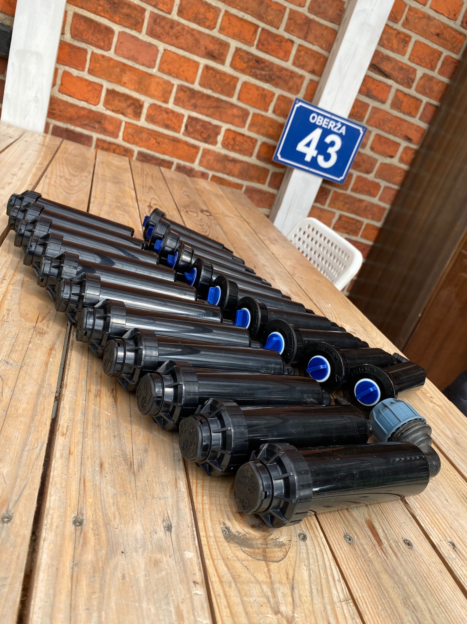 Rząd czarnych zraszaczy ogrodowych na drewnianym stole, z ceglanym murem i tabliczką z numerem domu w tle. Widok z góry, ostre światło.