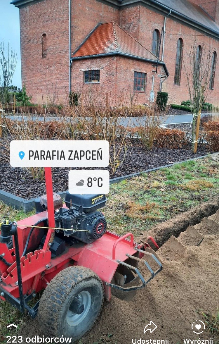 Czerwona glebogryzarka wykopuje wąski rów w ziemi, prawdopodobnie pod instalację nawadniającą, z widocznym kościołem w tle.