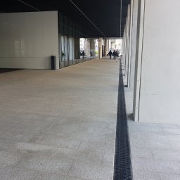 K&K KRZYSZTOF KAZIÓW - Szeroki chodnik wyłożony szarą kostką z odpływem liniowym, prowadzący wzdłuż budynku z przeszkloną fasadą i obrotowymi drzwiami wejściowymi, z przechodzącymi ludźmi w tle.