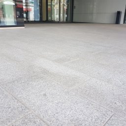 K&K KRZYSZTOF KAZIÓW - Jasnoszara kostka granitowa ułożona na zewnątrz budynku z dużymi przeszkleniami, widoczna spoinowania między kostkami.