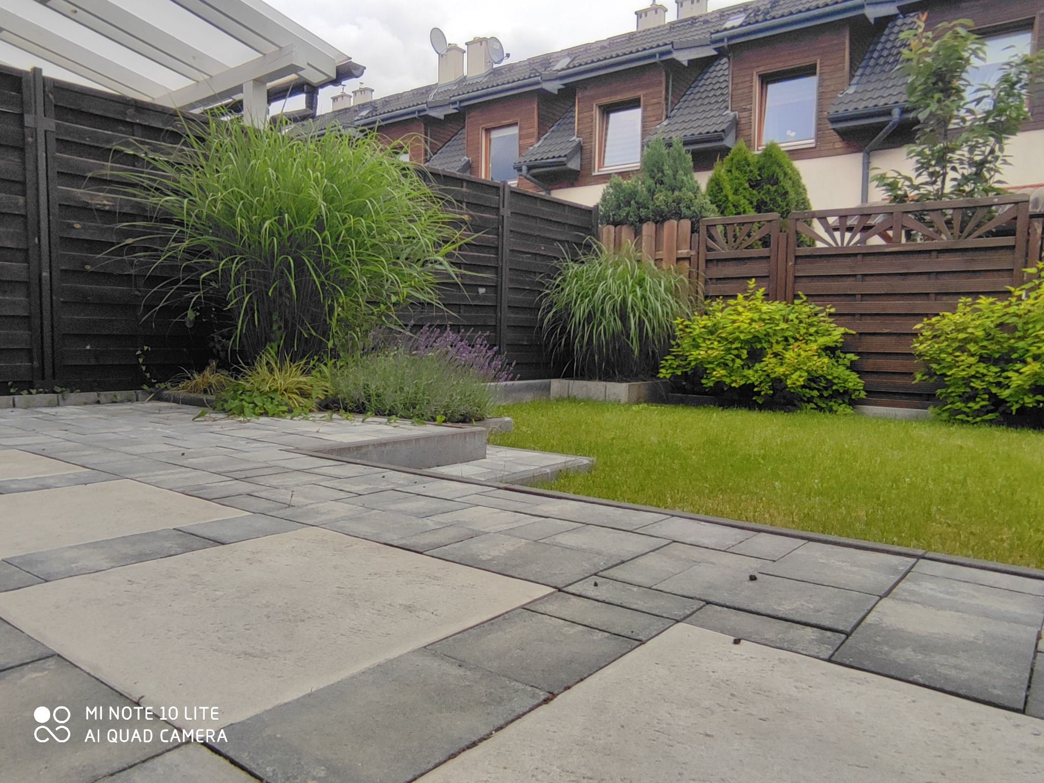 Ułożona nawierzchnia z kostki betonowej w odcieniach szarości i beżu, obrzeża z ciemniejszej kostki, w tle trawnik, roślinność ozdobna i drewniane ogrodzenie.