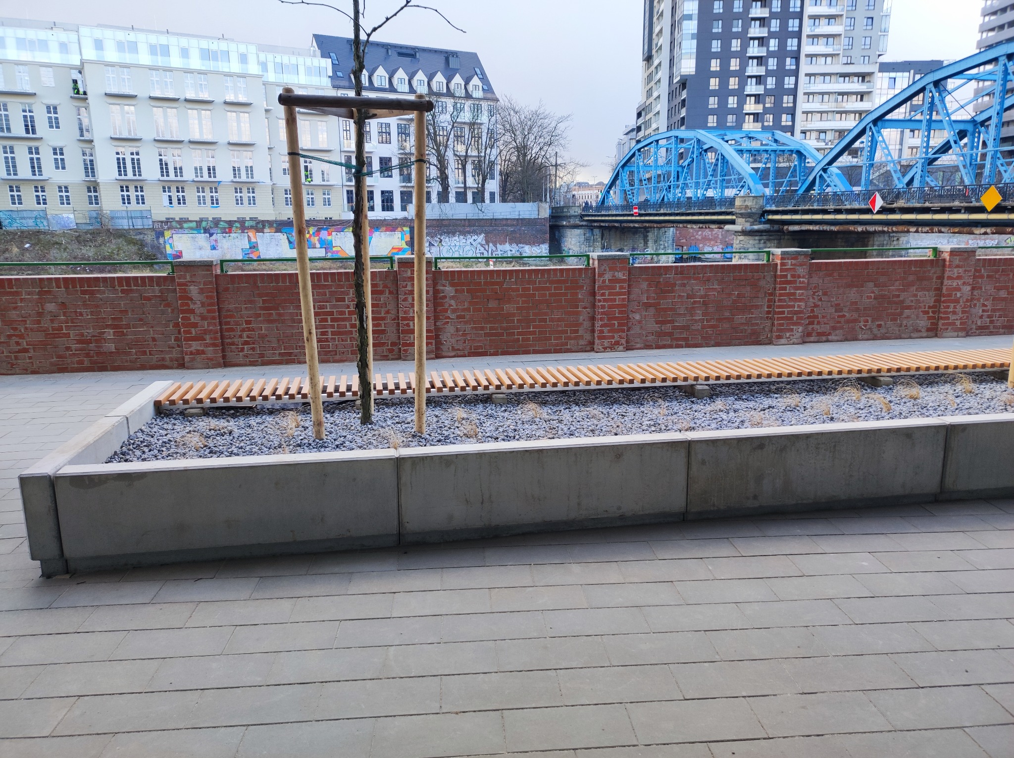 Miejska przestrzeń z betonową donicą wypełnioną drobnym żwirem, drewnianymi listwami i młodymi drzewkami, w tle ceglany mur, budynki i błękitny most.