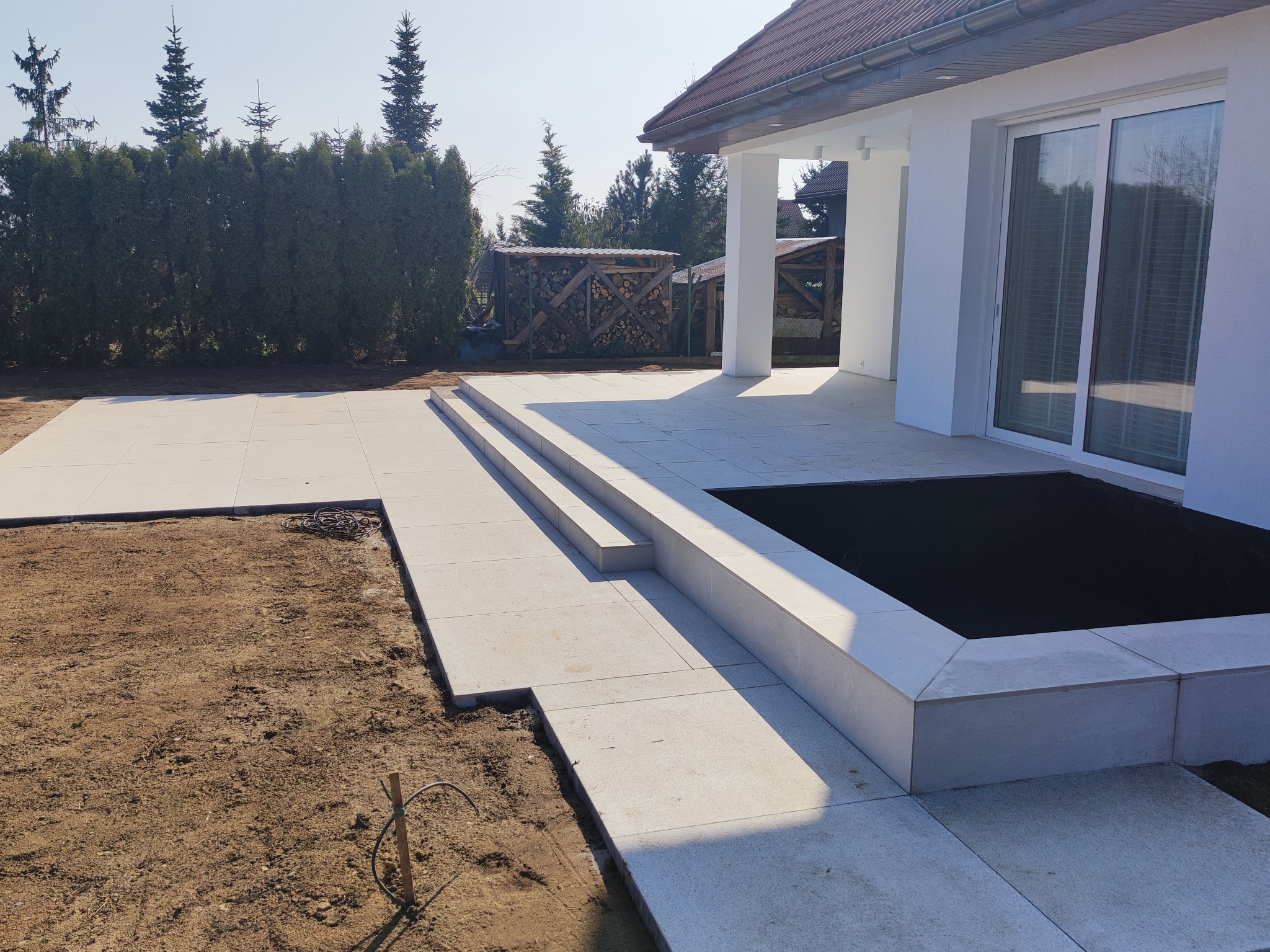 Jasnoszara nawierzchnia tarasu z płyt betonowych w trakcie układania, widoczne schody i rabata, w tle dom z białymi ścianami i sterta drewna.