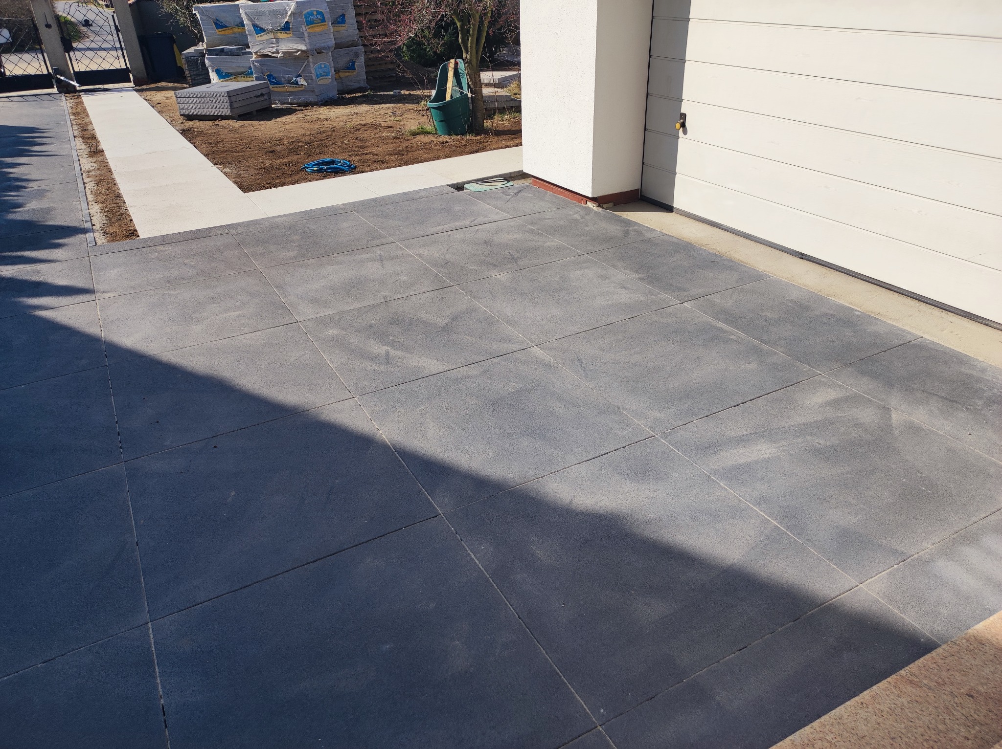 Szara nawierzchnia z płyt betonowych przed garażem, w tle ułożone palety z materiałami budowlanymi i brama wjazdowa.