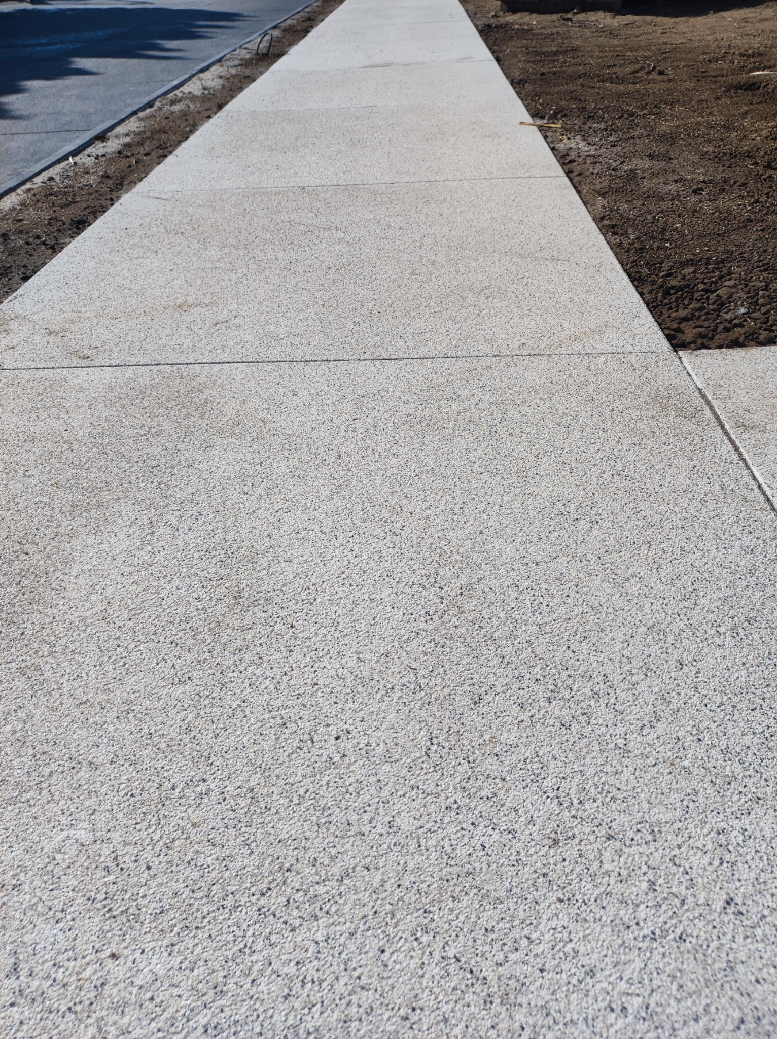 Nowa, jasnoszara nawierzchnia z kostki betonowej o drobnej fakturze, ułożona wzdłuż chodnika z widoczną świeżą ziemią po bokach.