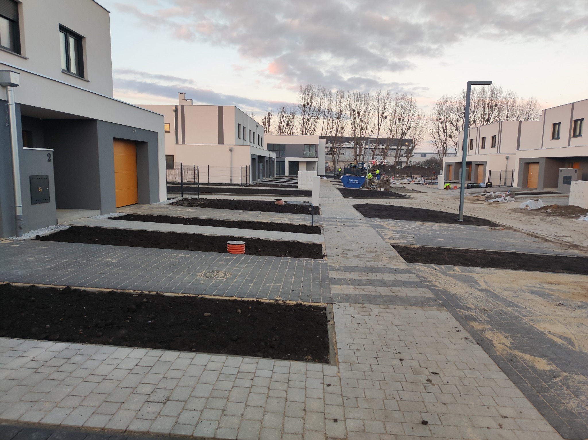 Nowoczesne osiedle w trakcie budowy z ułożoną szarą kostką brukową i przygotowanymi rabatami ziemnymi, w tle widoczny sprzęt budowlany i pracownicy.
