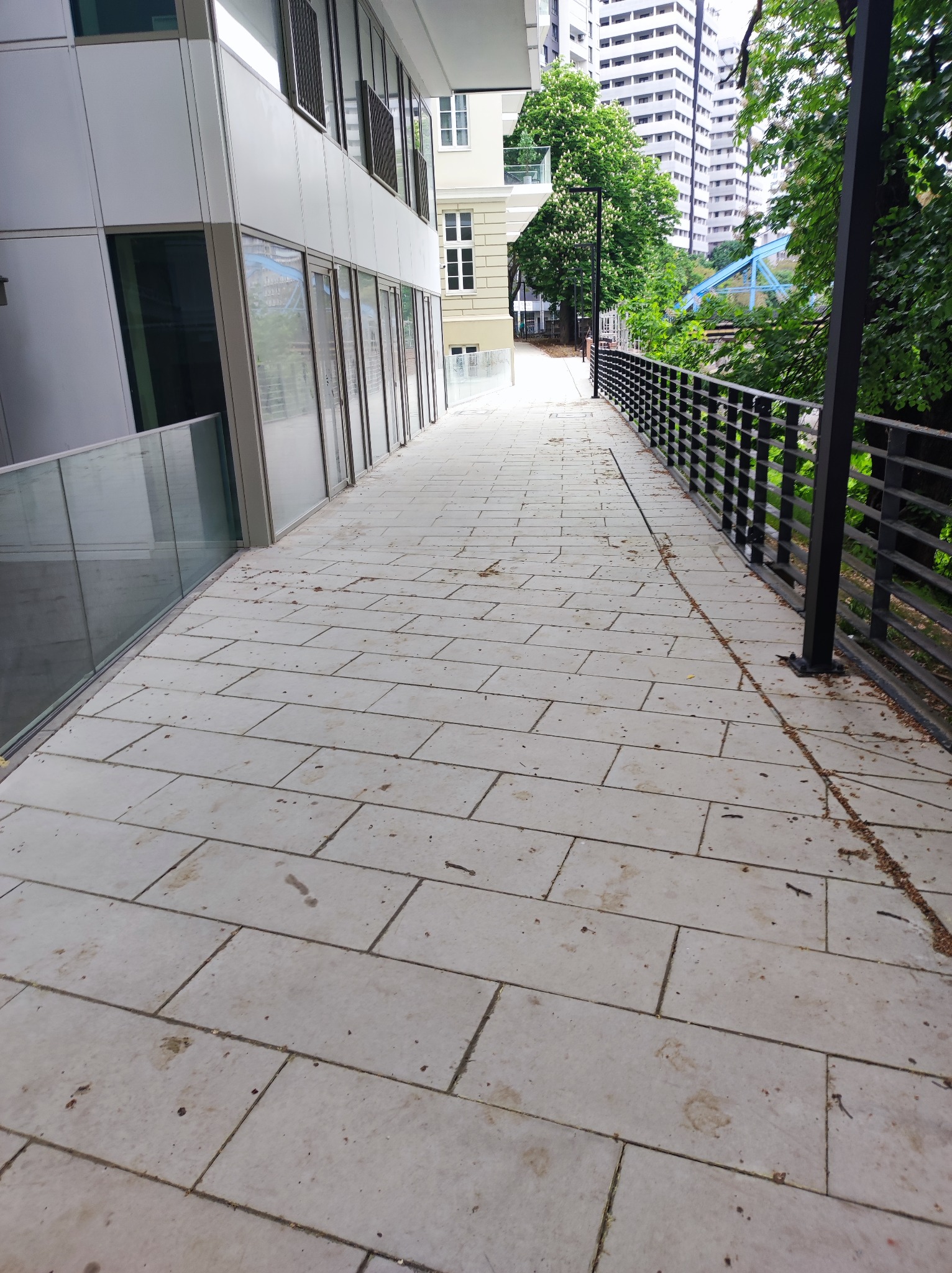 Wyłożona jasnymi płytami chodnikowa ścieżka biegnie wzdłuż nowoczesnego budynku z przeszklonymi ścianami i metalową balustradą, w tle widoczne drzewa i wysokie budynki.
