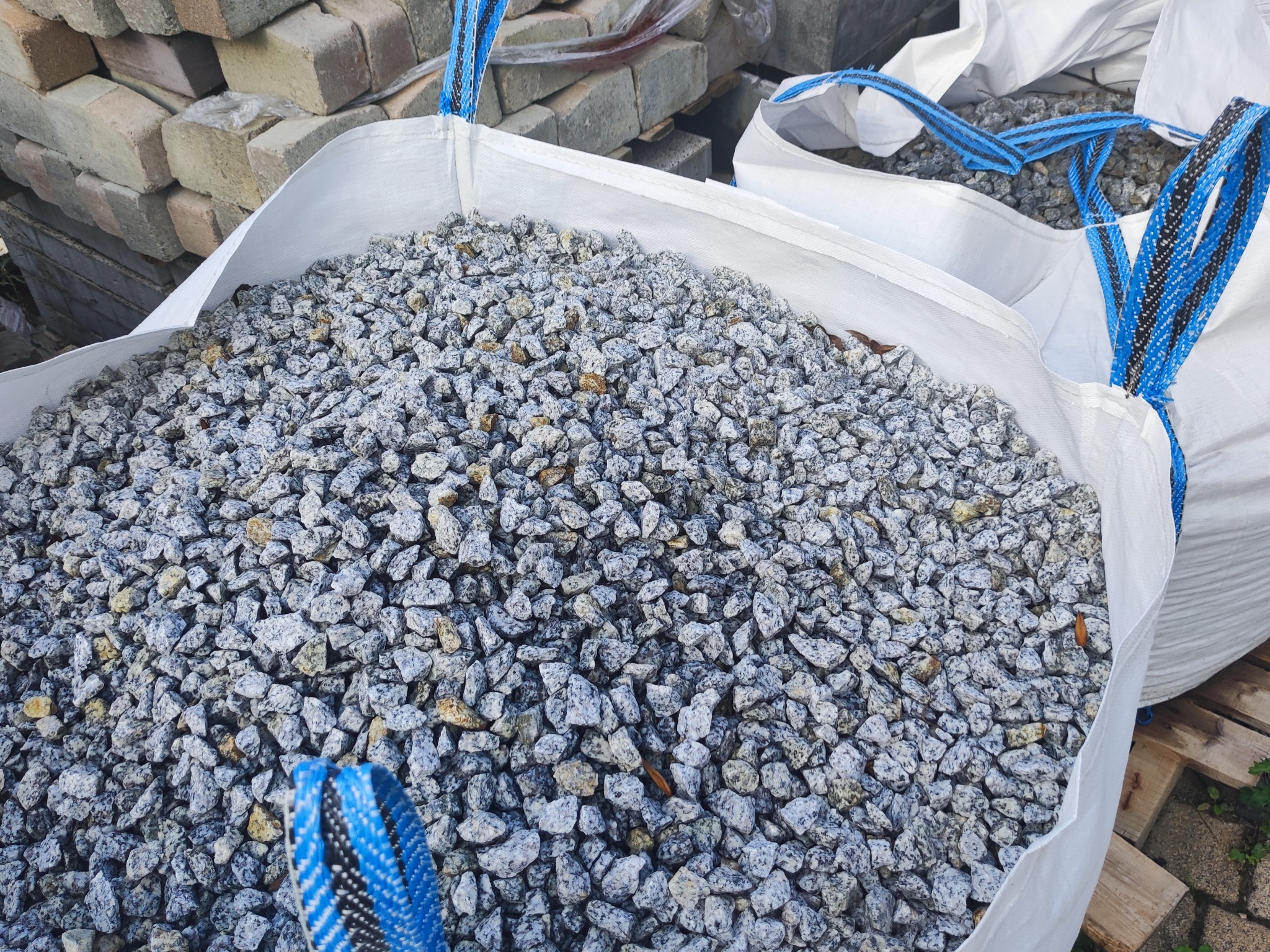 Worek typu Big Bag wypełniony ozdobnym, szaro-białym żwirem granitowym, widoczne niebieskie uchwyty worka i paleta w tle.
