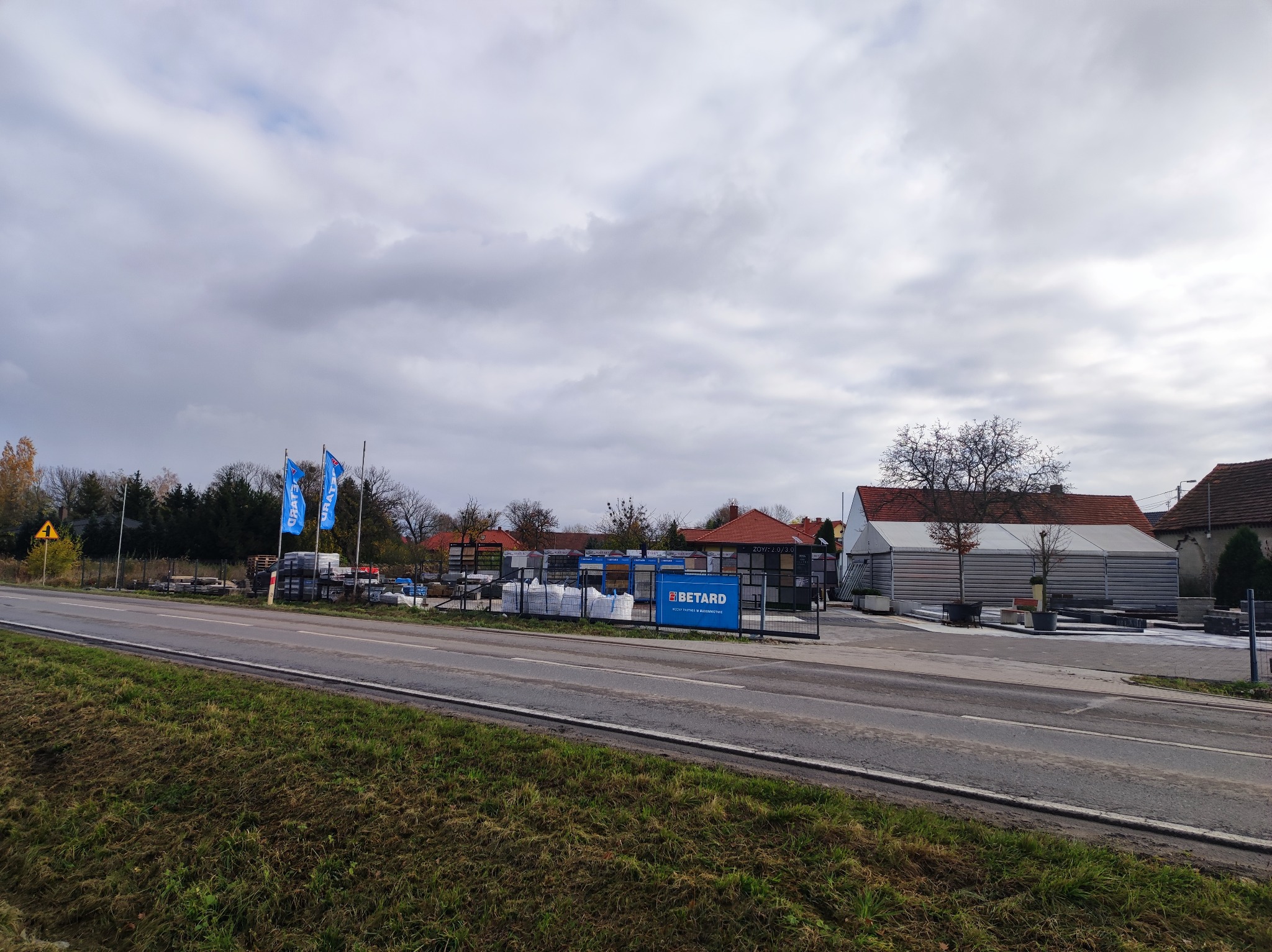Widok na skład budowlany Betard z ulicy, z widocznymi materiałami budowlanymi, flagami firmowymi i ekspozycją produktów na zewnątrz.