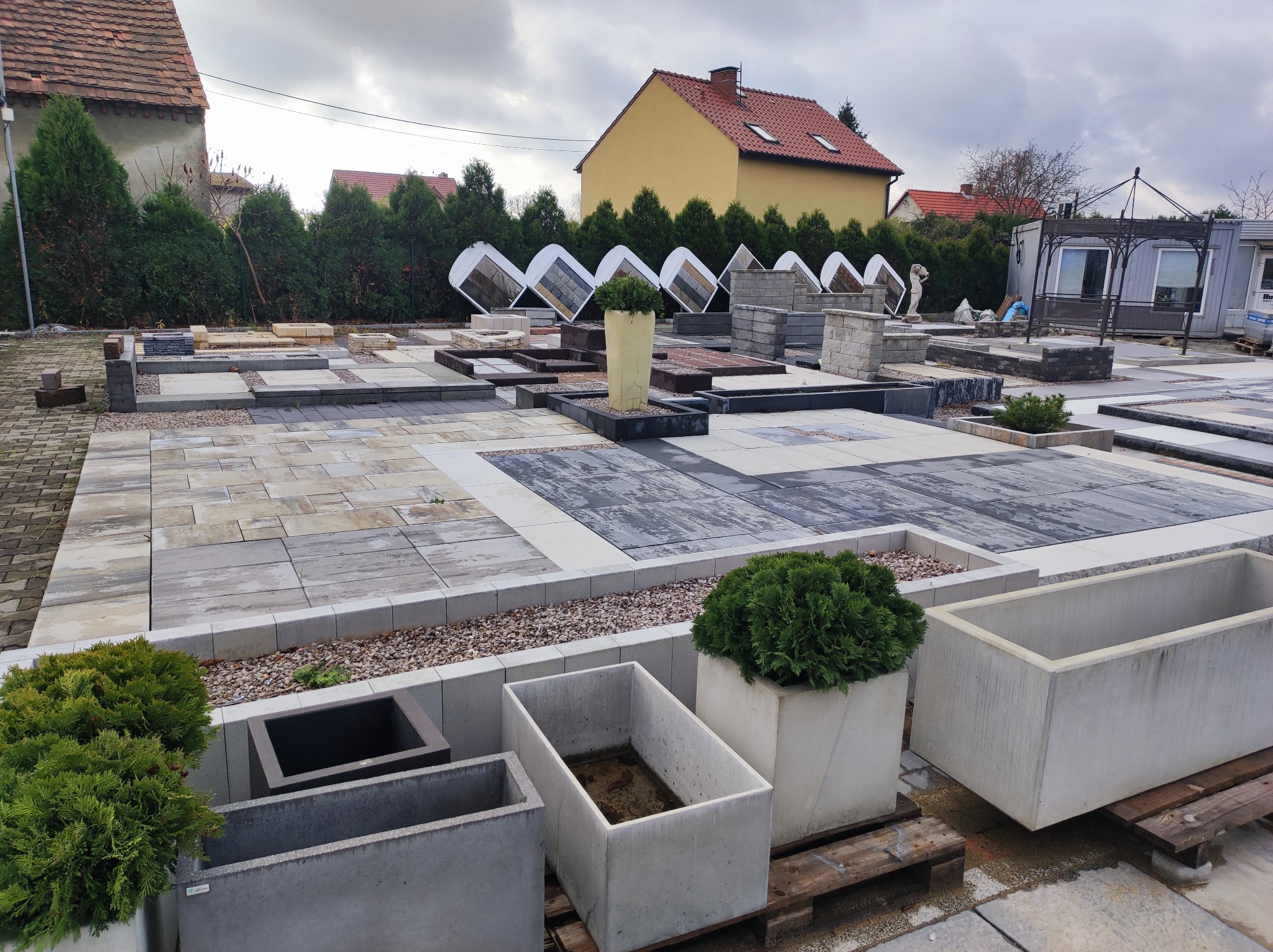 Ekspozycja różnorodnych wzorów kostki brukowej i betonowej w Cesarzowicach, z donicami i roślinami, prezentowana na zewnątrz w pochmurny dzień.