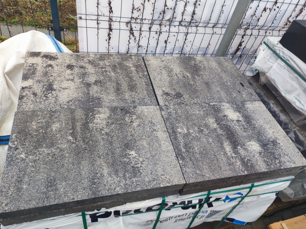Cztery szare płyty chodnikowe o porowatej fakturze, ułożone na palecie z folią, na tle metalowego ogrodzenia i roślinności.