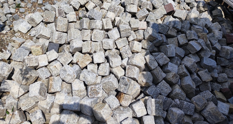 Stos szarej kostki o nieregularnych kształtach, widoczne drobne ziarna i naturalne przebarwienia kamienia, częściowo oświetlone słońcem, tworzące cienie.