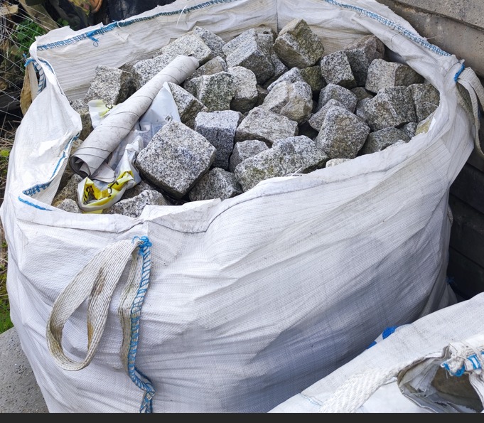 Worek z szarą kostką granitową w Cesarzowicach, widoczne nierównomierne bryły i zwinięty arkusz papieru na wierzchu.