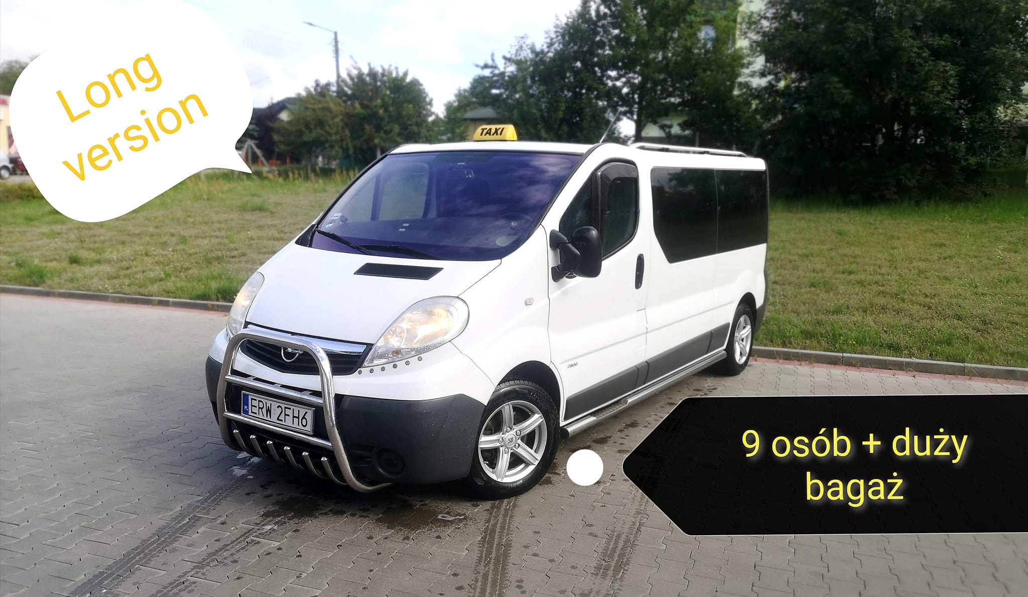 Biały, długi bus taxi z chromowanym orurowaniem z przodu, zaparkowany na kostce brukowej przed zielonym trawnikiem.