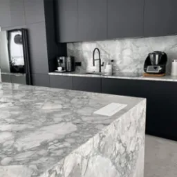 Nowoczesna kuchnia z czarnymi szafkami i wyspą z blatem z szarego marmuru Carrara, dopasowanym do ściany nad blatem kuchennym. Na blacie ekspres do kawy i Thermomix.