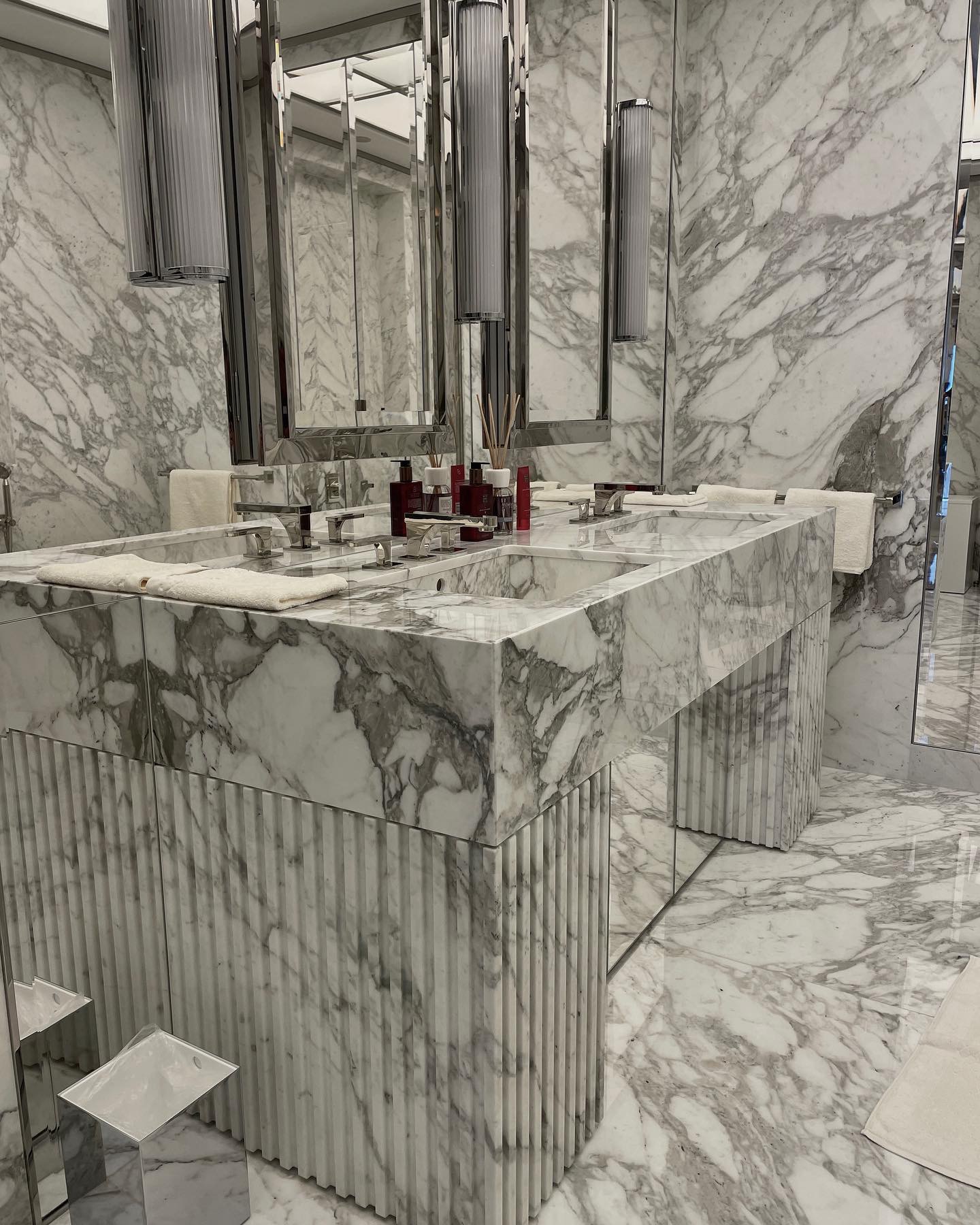 Elegancka łazienka wykończona białym marmurem, z podwójną umywalką i lustrami, oświetlona wiszącymi lampami, z widocznymi akcesoriami łazienkowymi i ręcznikami.