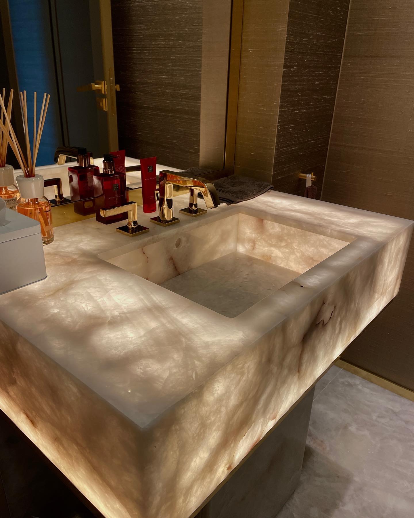 Oświetlony od spodu, marmurowy blat umywalkowy z wbudowanym zlewem i złotą armaturą, uzupełniony o kosmetyki i dyfuzor zapachowy.