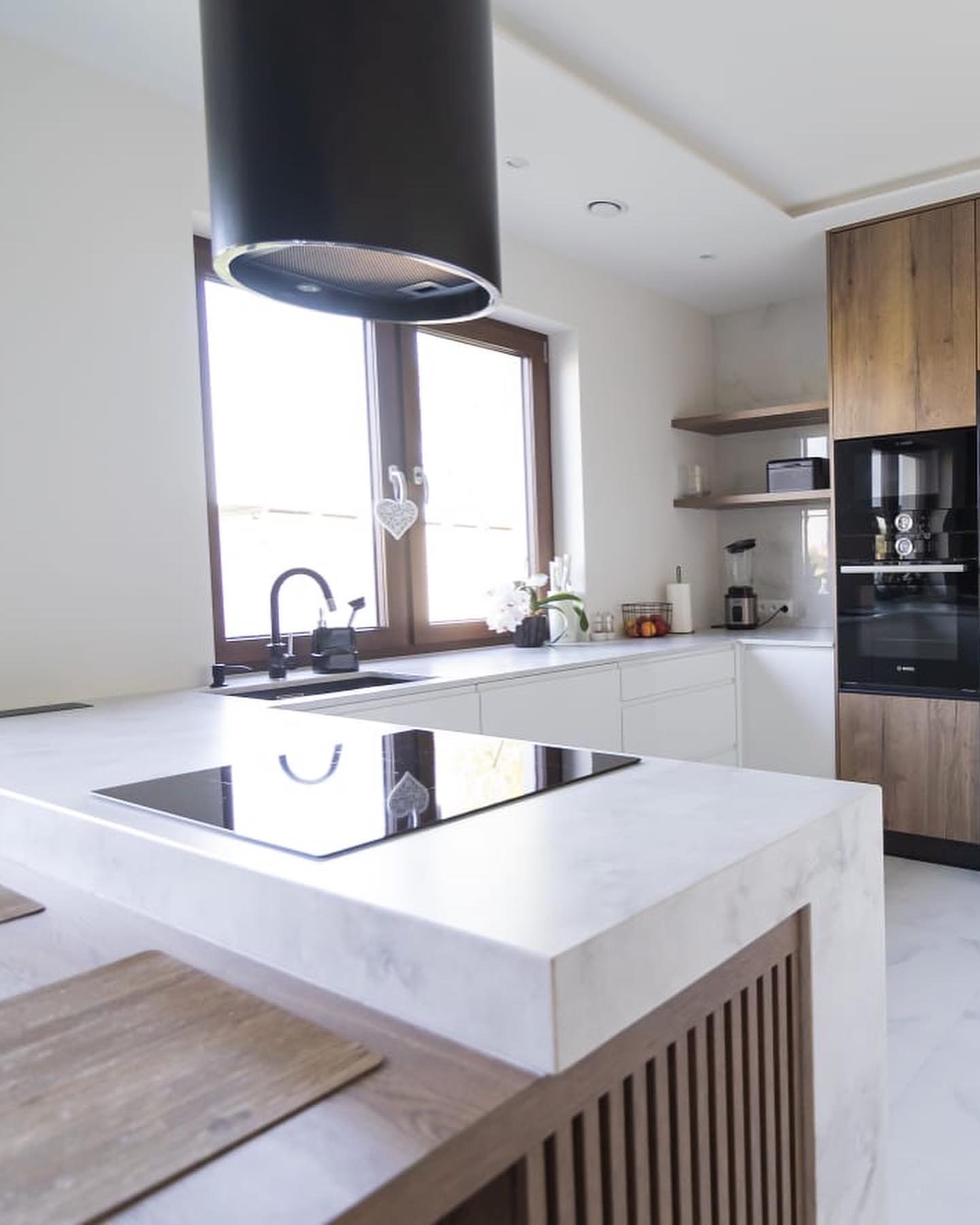 Biały blat z marmurowym wzorem, wbudowana czarna płyta indukcyjna, nowoczesna kuchnia z drewnianymi szafkami i czarnym okapem cylindrycznym.