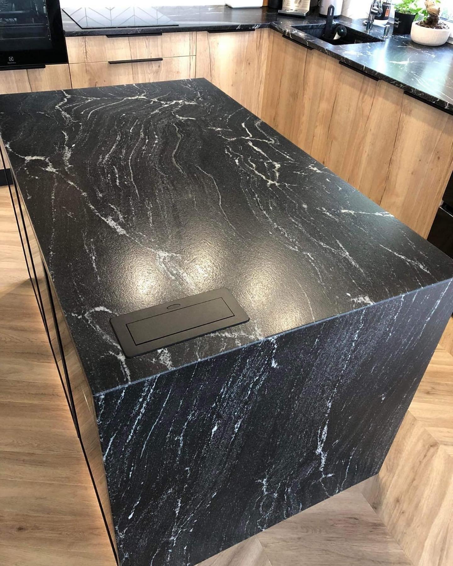 Czarny, polerowany blat z kamienia naturalnego z białymi żyłkami na wyspie kuchennej, z wbudowanym gniazdem elektrycznym, na tle drewnianych szafek i parkietu.