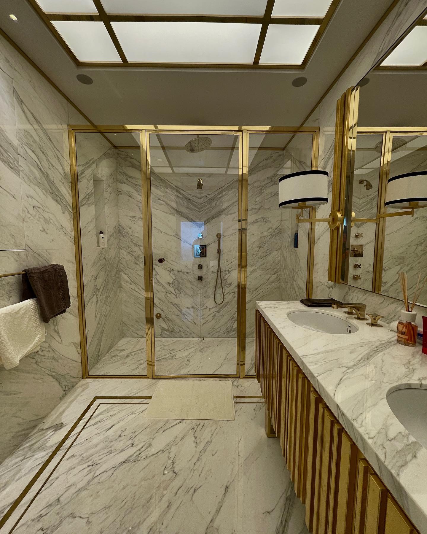 Luksusowa łazienka wykończona białym marmurem z żyłami, z kabiną prysznicową typu walk-in z złotymi profilami i armaturą, oraz podwójną umywalką z blatem z tego samego marmuru i szafką z pionowymi,...