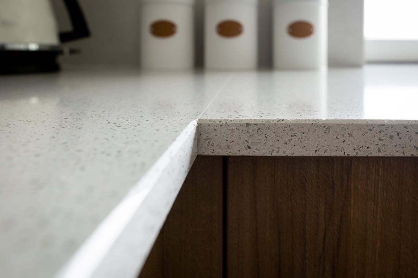 Jasny blat kuchenny z konglomeratu kwarcowego, widoczne krawędzie i fragment szafki pod zlewem, w tle pojemniki na przyprawy.