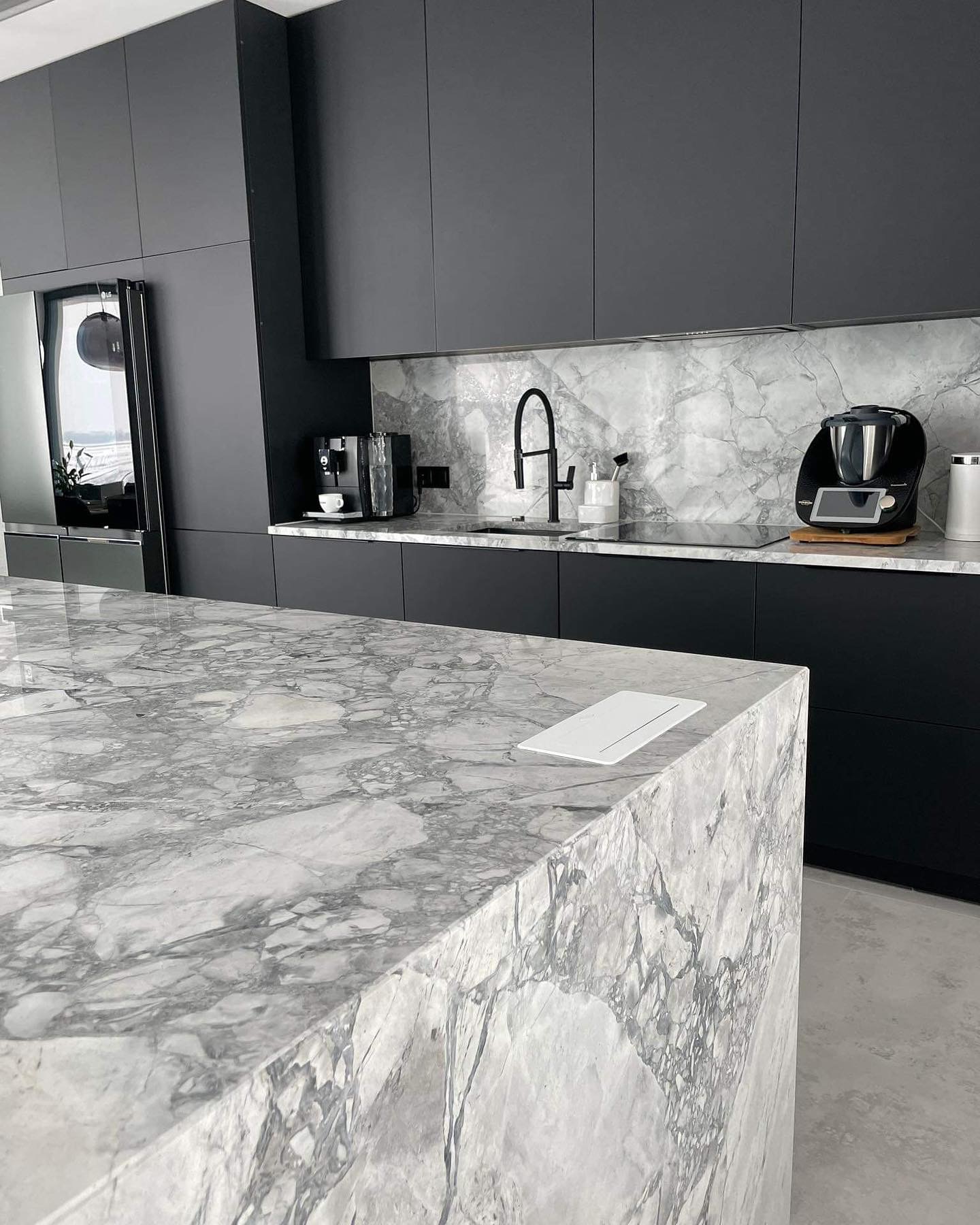Nowoczesna kuchnia z czarnymi szafkami i wyspą z blatem z szarego marmuru Carrara, dopasowanym do ściany nad blatem kuchennym. Na blacie ekspres do kawy i Thermomix.