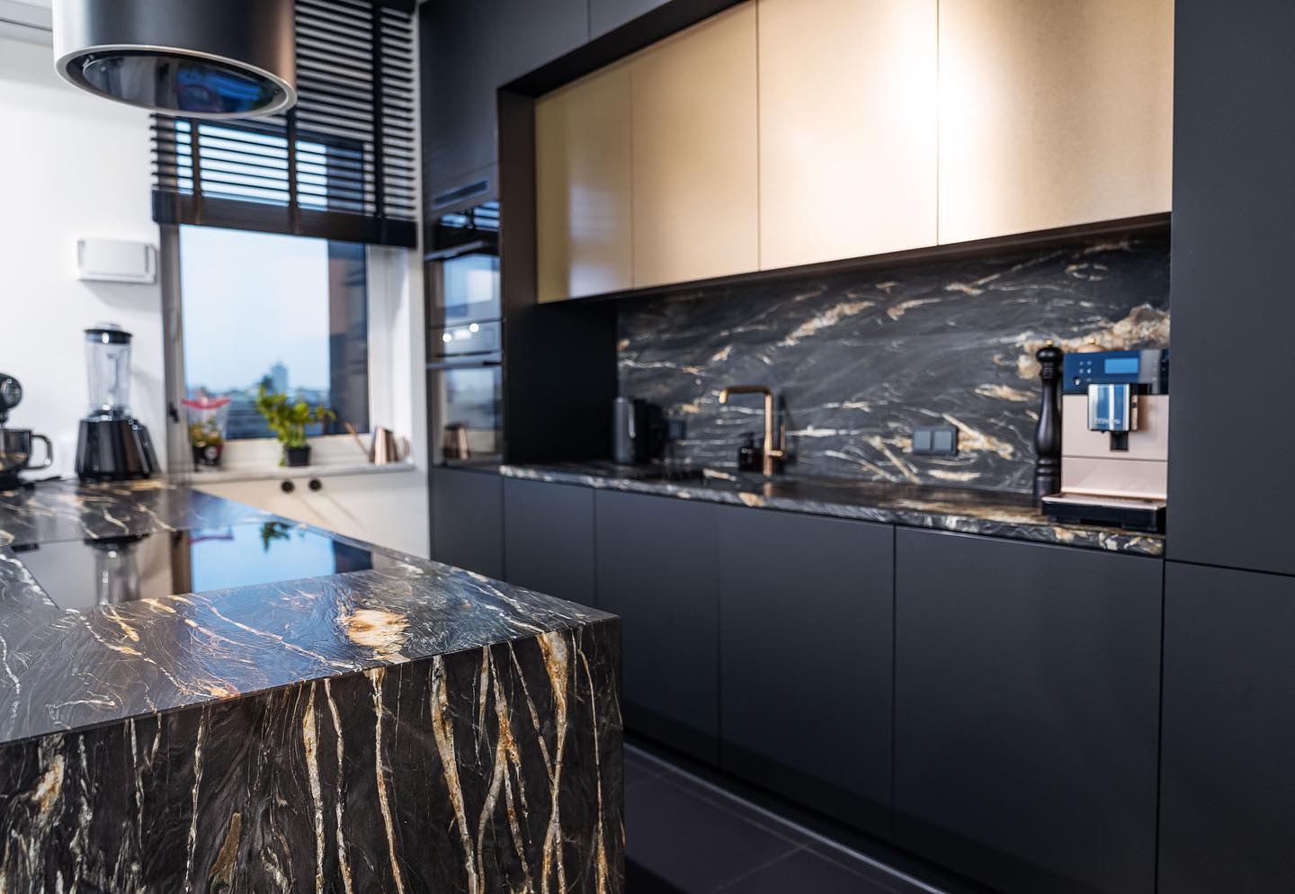 Nowoczesna kuchnia z czarnymi szafkami i blatem z czarnego kamienia z wyraźnym złotym użyleniem, widoczny ekspres do kawy i bateria w kolorze miedzi.