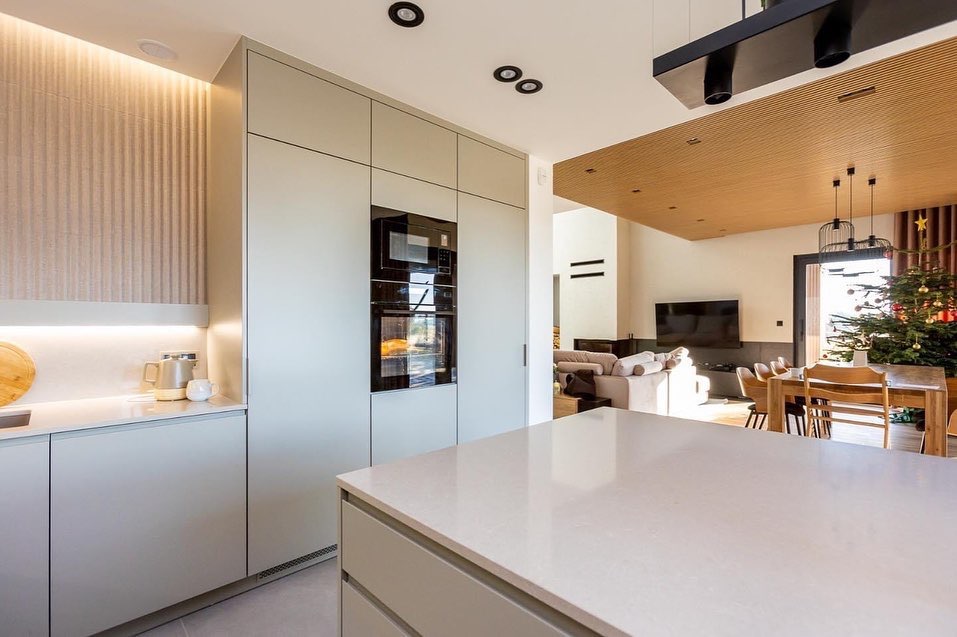 Nowoczesna kuchnia z wyspą i blatem o jasnej, jednolitej barwie; w tle salon z kominkiem i choinką.