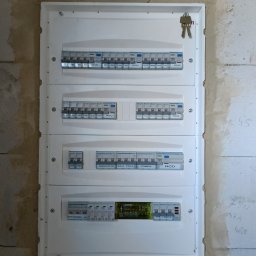 Rozdzielnica elektryczna Hager z bezpiecznikami i opisami obwodów (indukcja, siła, pompa) zamontowana na surowej ścianie z betonu komórkowego. Klucze wiszą na górze.