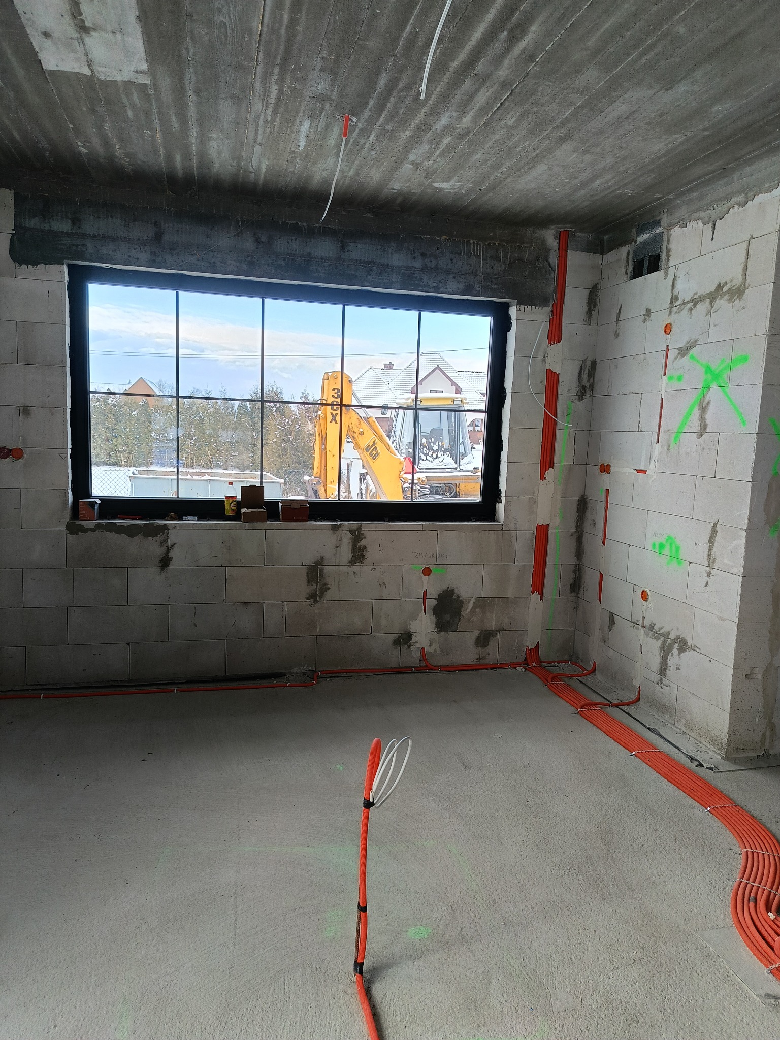 Wnętrze w trakcie prac wykończeniowych z widoczną instalacją elektryczną w peszlach na ścianach z betonu komórkowego. W oknie widoczny krajobraz z koparką.