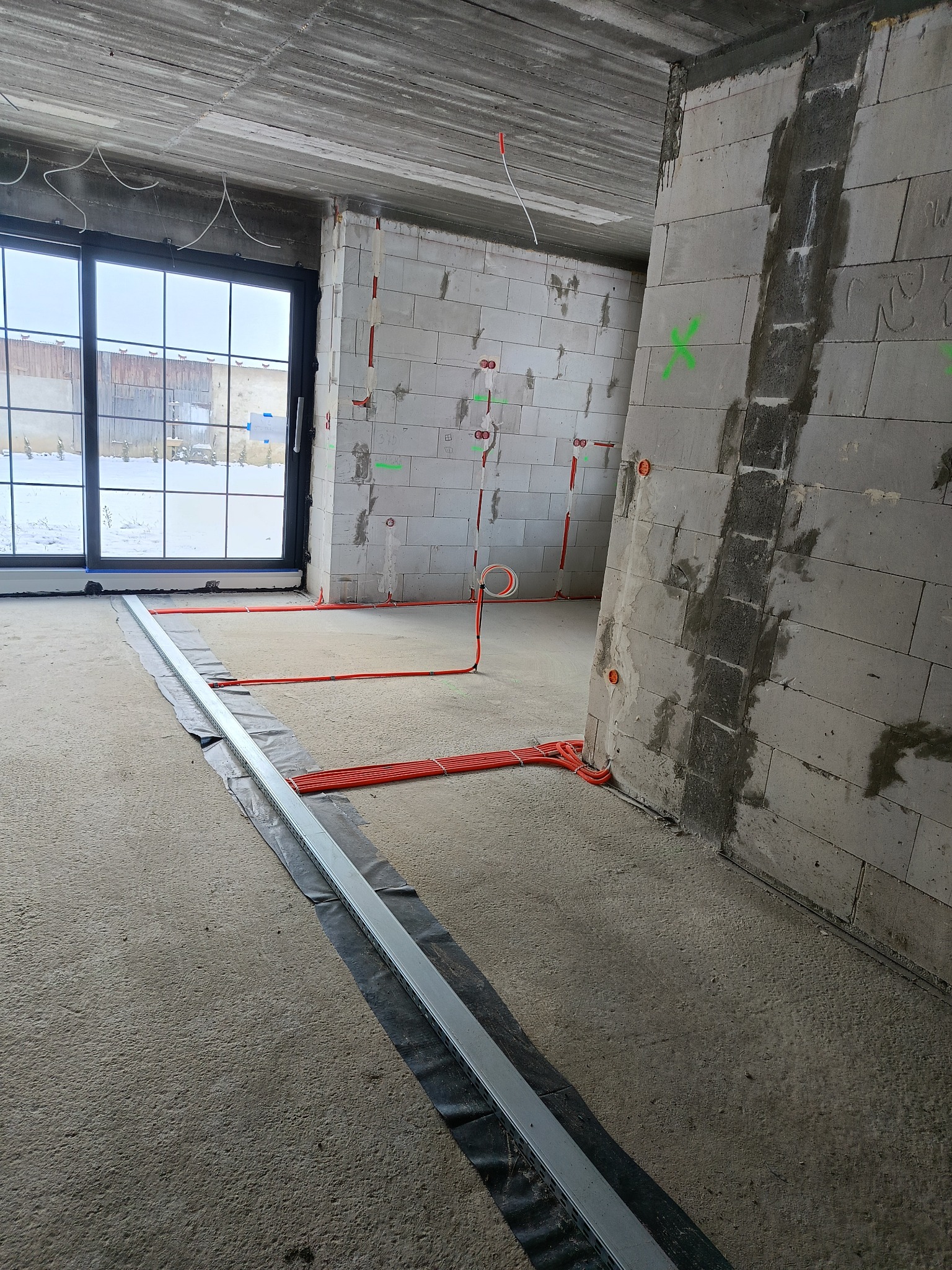 Wnętrze w trakcie prac: widoczne przewody elektryczne w peszlach na ścianach z betonu komórkowego i w korycie podłogowym, okno z widokiem na zimowy krajobraz.