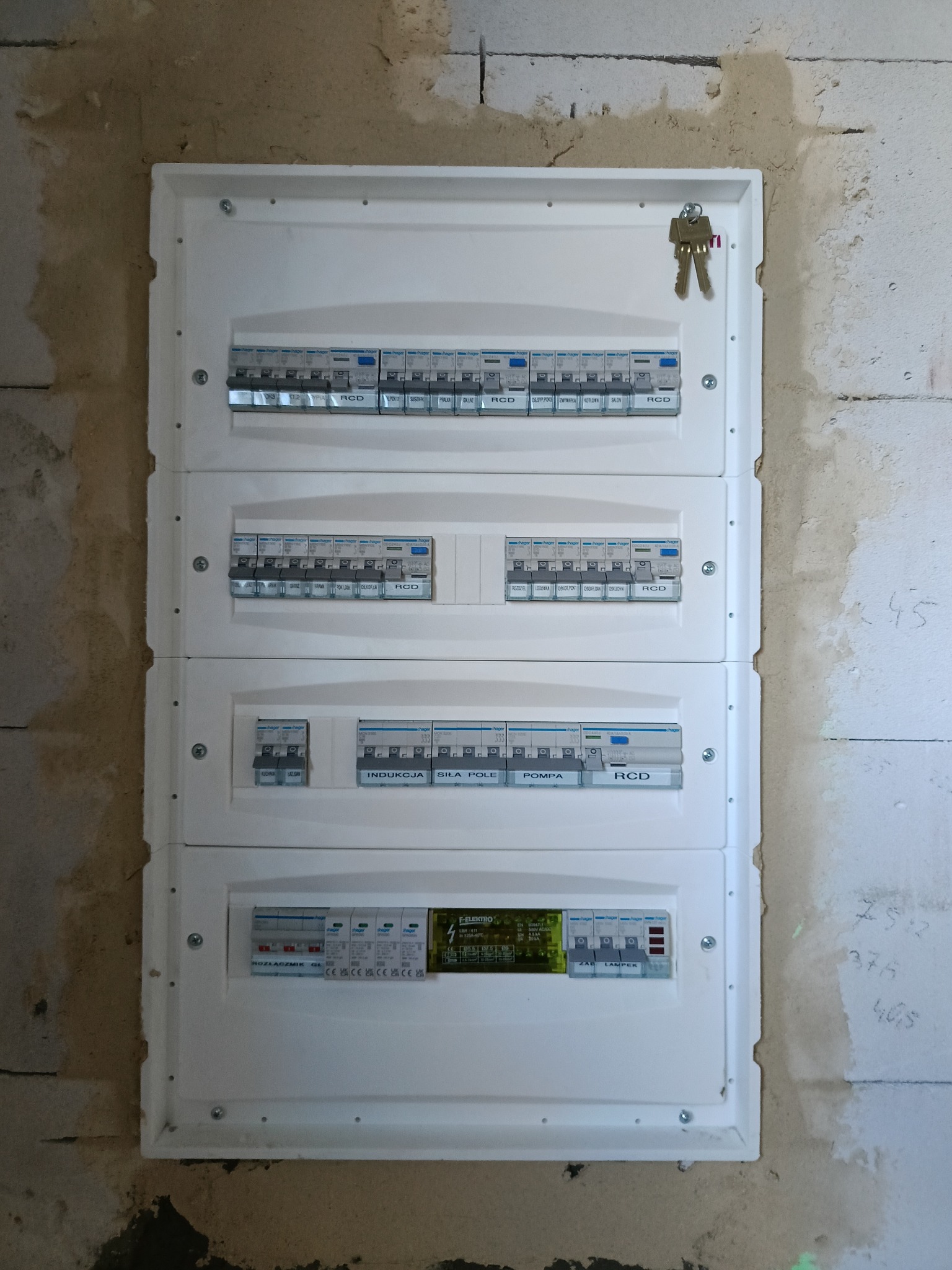Rozdzielnica elektryczna Hager z bezpiecznikami i opisami obwodów (indukcja, siła, pompa) zamontowana na surowej ścianie z betonu komórkowego. Klucze wiszą na górze.