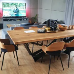 Stół z blatem z litego drewna o nieregularnych krawędziach, wsparty na czarnej, metalowej konstrukcji, otoczony krzesłami z drewnianym oparciem i czarnym siedziskiem, w tle salon z szarą sofą...