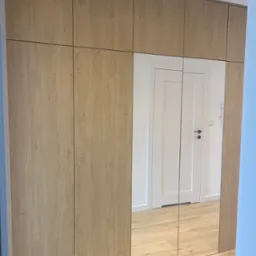 Wysoka zabudowa z drewnopodobnych paneli, wbudowane lustrzane drzwi odbijają wnętrze z białą ścianą i drzwiami.